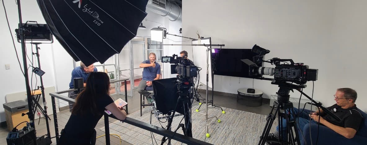 A two-camera interview set: crew operating Sony cinema cameras and a large softbox while the subject gives a thumbs-up from his seat
