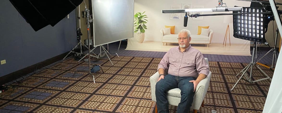 An interview subject seated in an armchair in front of a printed living-room backdrop, with a boom microphone, softbox and reflector set around him