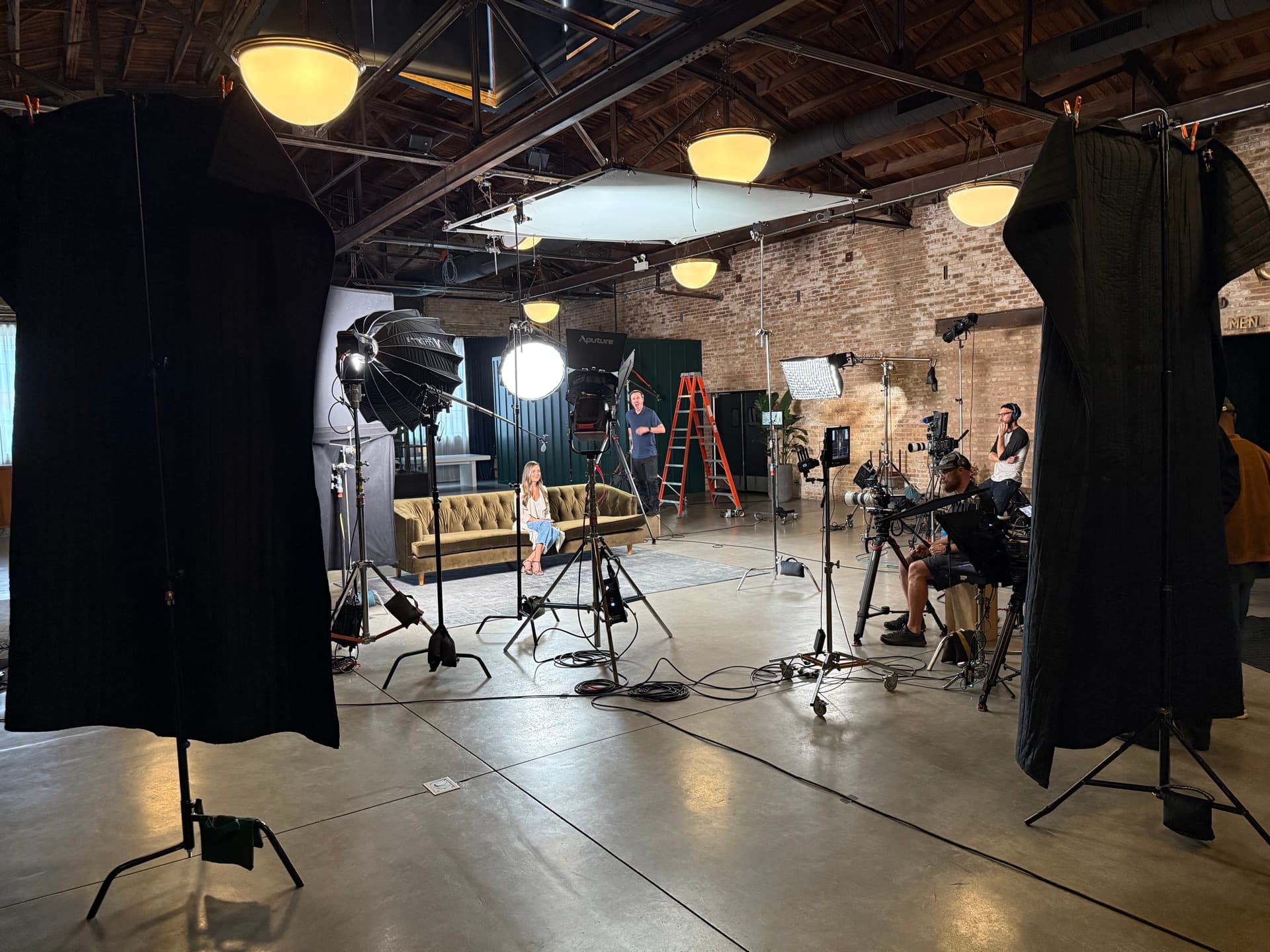 Lighting, camera and sound crew set up around a velvet-couch conversation set in a brick-walled loft